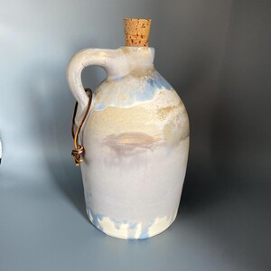 May include: A handmade ceramic jug with a handle and a cork stopper. The jug has a light blue, beige, and white glaze. A brown leather strap is attached to the handle. The jug is approximately 25 cm tall.