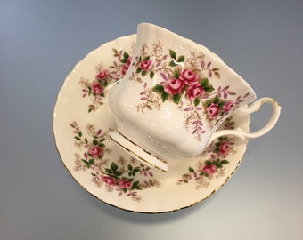 May include: A white teacup and saucer with a floral pattern. The cup and saucer are decorated with pink roses and green leaves. The edges of the cup and saucer are trimmed with gold.