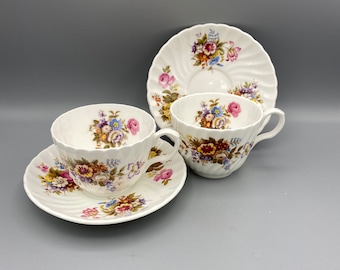 Puede incluir: Dos tazas de té blancas con diseños florales y platillos a juego. Las tazas tienen un borde festoneado y un diseño floral en tonos de rosa, azul, amarillo y verde. Los platillos tienen un diseño floral similar y un borde festoneado.