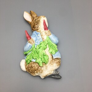 Puede incluir: Figura de cerámica de un conejo con una chaqueta azul, sosteniendo zanahorias y hojas verdes. El conejo tiene pelaje marrón y detalles blancos. La figura está pintada con un acabado brillante. El conejo está sentado.