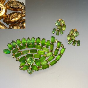 Pode incluir: Um conjunto de broche e brincos de vidro verde e tom dourado. O broche tem a forma de uma folha e é feito de muitas pequenas pedras de vidro verde. Os brincos também são feitos de pedras de vidro verde e têm a forma de folhas.