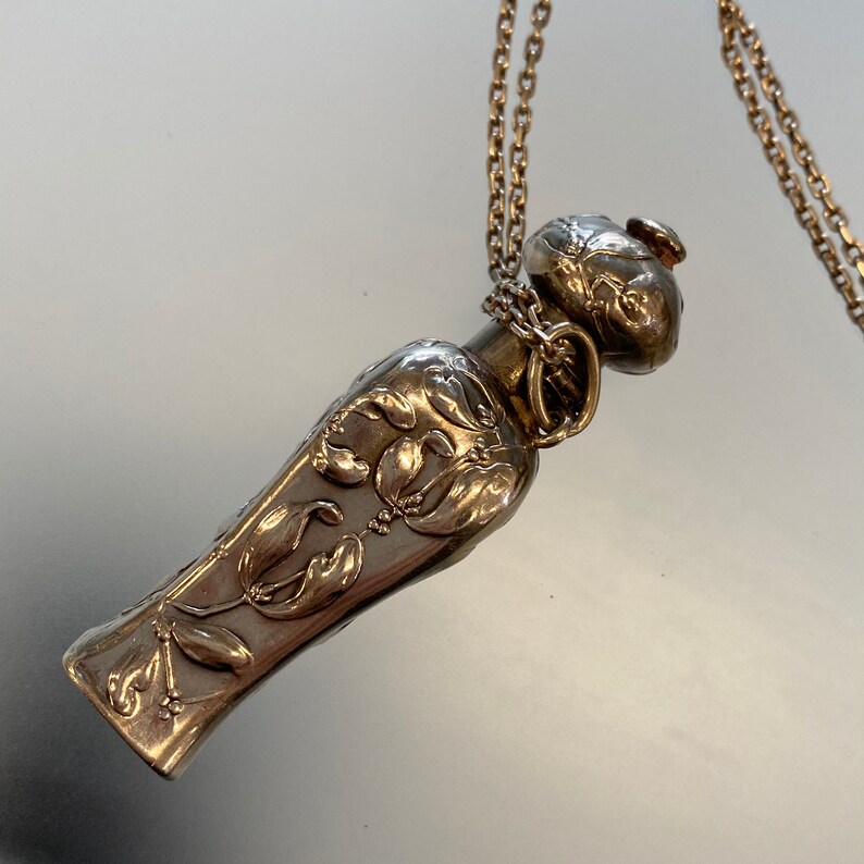 May include: A silver-toned pendant necklace featuring a decorative perfume bottle-shaped charm. The charm is adorned with floral relief patterns and hangs from a delicate chain. The design is detailed and elegant, suitable for a vintage or antique jewelry collection.
