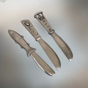 May include: Three silver butter knives with decorative handles. One knife features a fish design, another has a floral motif, and the third has a geometric pattern. The knives are arranged on a reflective surface.
