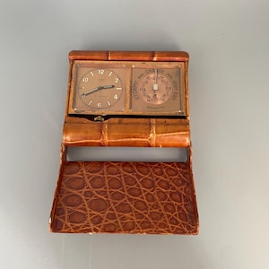 May include: A vintage travel clock and barometer in a brown leather case. The clock face has Roman numerals and the barometer has a needle that points to "Change".