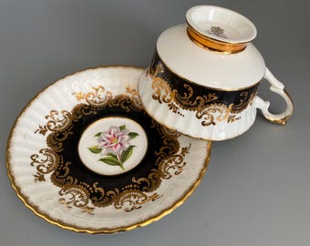May include: A white teacup and saucer set with gold trim and black and gold filigree designs. The saucer features a central floral design with pink and green petals. The teacup has a gold rim and handle.