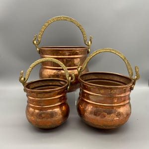 Puede incluir: Tres ollas de cobre con asas de latón. Las ollas son de diferentes tamaños, con una textura martillada en el cobre. Las asas arqueadas tienen detalles decorativos. Las ollas están dispuestas sobre un fondo gris.