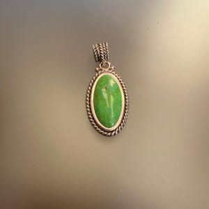 Pode incluir: Um pingente de prata com uma pedra oval de turquesa verde engastada em uma borda de corda.