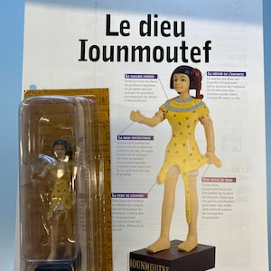 May include: A figurine of the Egyptian god Iounmoutef, standing on a brown base. The figurine is made of painted resin and depicts the god with a yellow tunic, a leopard skin, and a protective hand gesture. The figurine is packaged in a clear plastic box with the name "Iounmoutef" printed on it.