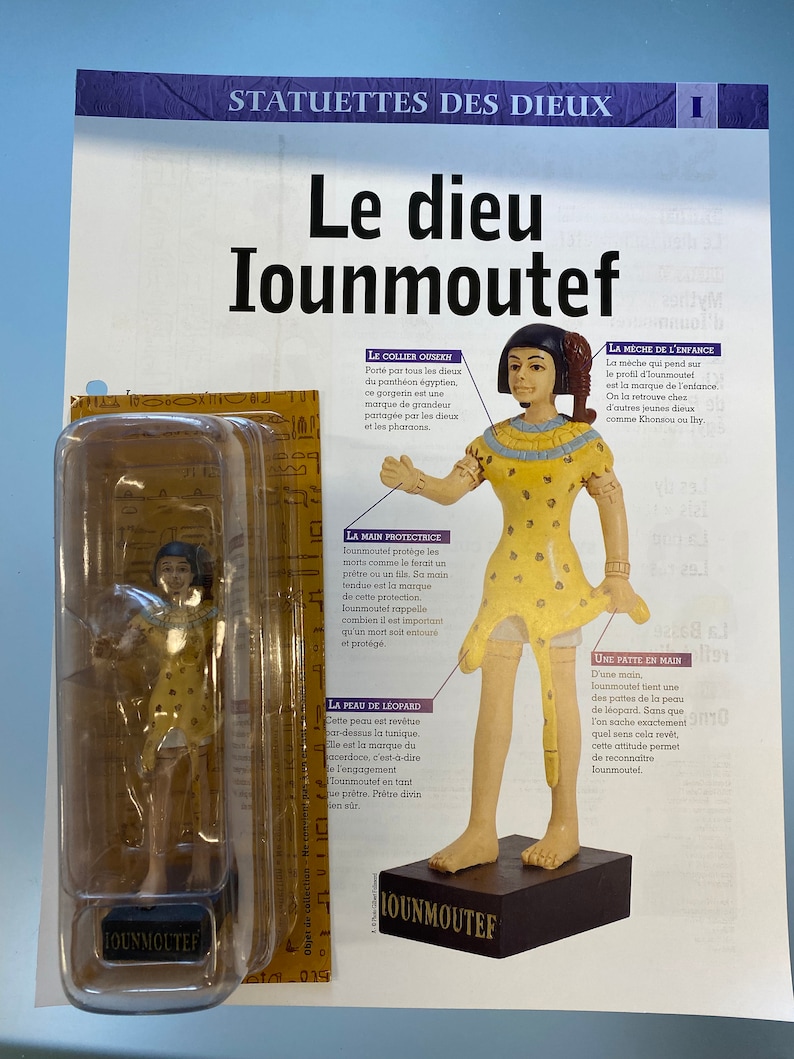 May include: A figurine of the Egyptian god Iounmoutef, standing on a brown base. The figurine is made of painted resin and depicts the god with a yellow tunic, a leopard skin, and a protective hand gesture. The figurine is packaged in a clear plastic box with the name "Iounmoutef" printed on it.