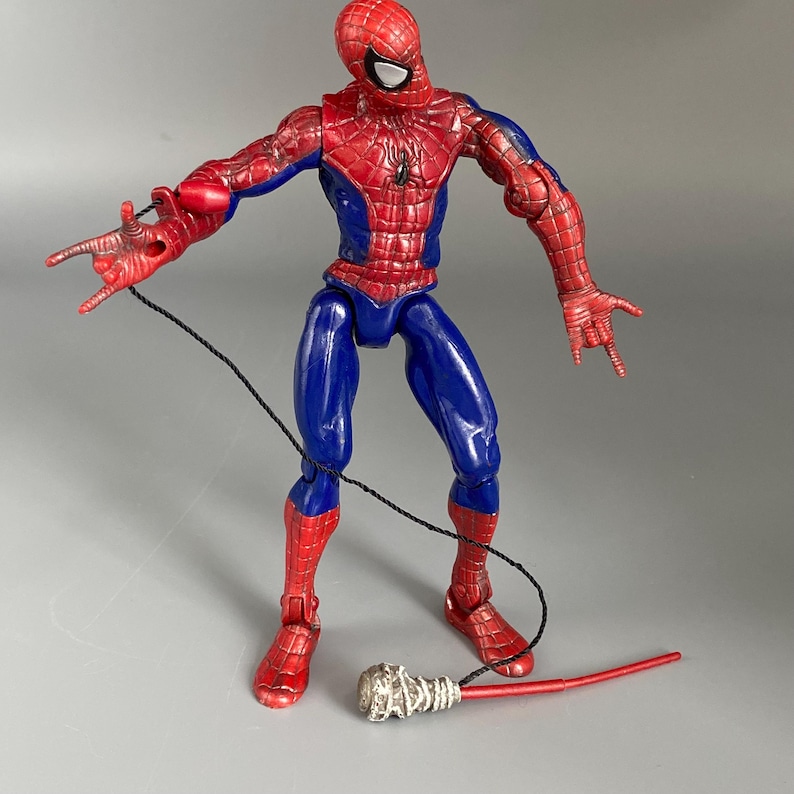 May include: A red and blue action figure of Spider-Man with a web shooter accessory. The figure is posed in a dynamic action pose with one arm extended and the other bent at the elbow. The figure has a detailed sculpt and paint job, including a spider logo on the chest.