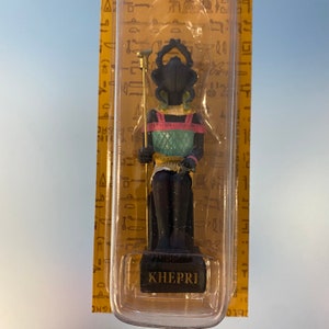 Peut inclure: Une petite figurine noire du dieu &eacute;gyptien Kh&eacute;pri, repr&eacute;sent&eacute; comme un scarab&eacute;e avec une t&ecirc;te humaine, portant une coiffe color&eacute;e et tenant un b&acirc;ton. La figurine est assise sur un tr&ocirc;ne et est expos&eacute;e dans un bo&icirc;tier en plastique transparent sur fond jaune. Le bo&icirc;tier porte l'inscription "KHEPRI" en bas.