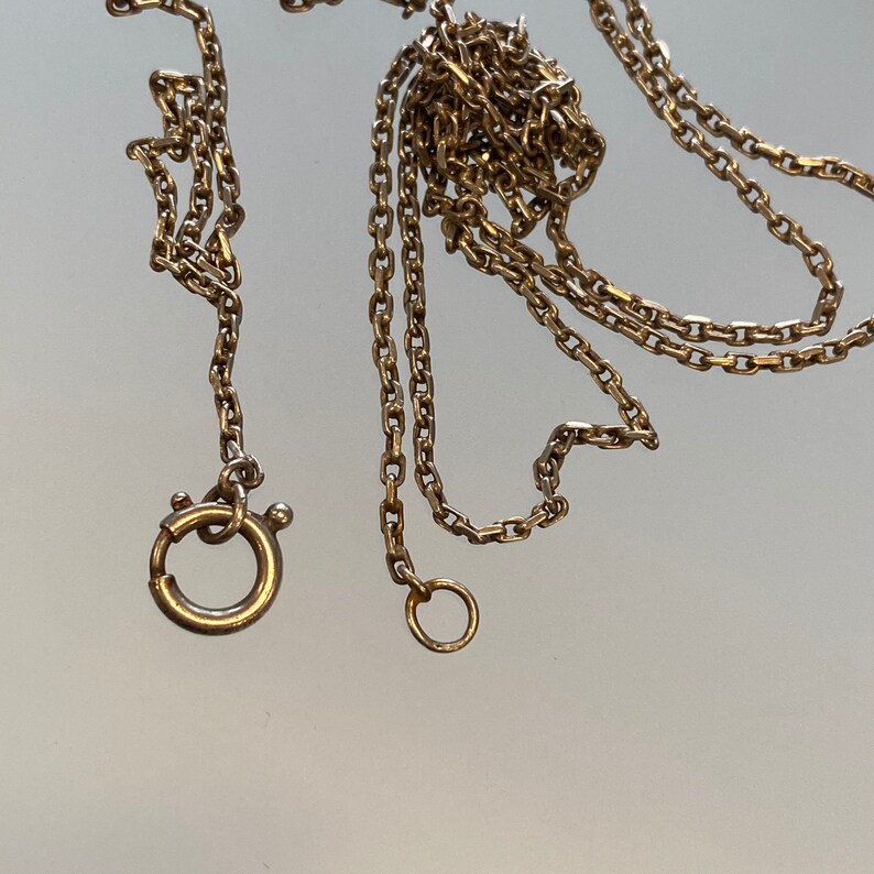 May include: Close-up of a gold-colored chain necklace with a spring ring clasp and a small circular ring. The chain is made of small, rectangular links. The necklace is displayed on a reflective surface.