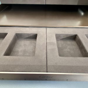 May include: A brown velvet lined jewelry box with four compartments. The box is open and the compartments are empty. The box has a silver metal trim and the word 'Cartier' is engraved on the front.