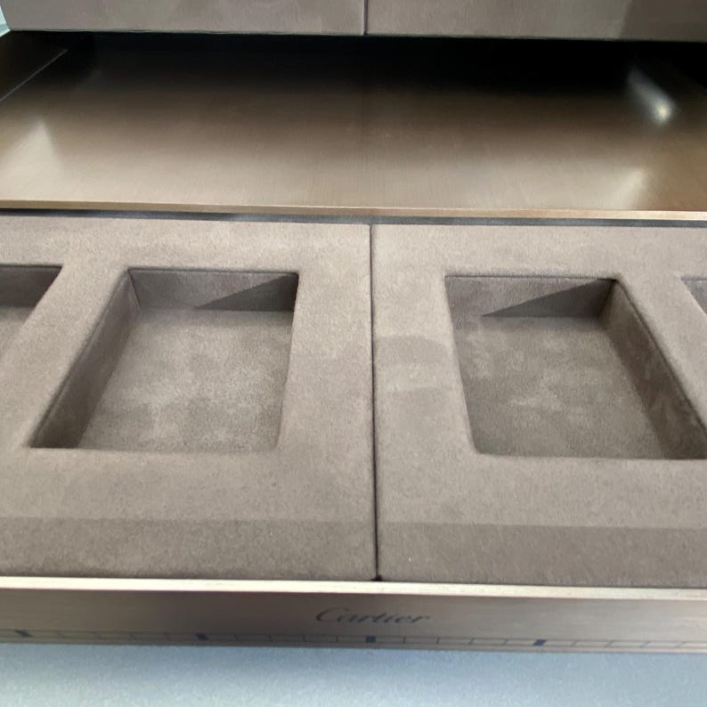 May include: A brown velvet lined jewelry box with four compartments. The box is open and the compartments are empty. The box has a silver metal trim and the word 'Cartier' is engraved on the front.
