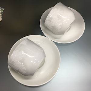 Peut inclure: Deux tasses à thé et soucoupes en céramique blanches. Les tasses ont un fond gris clair avec un motif de feuilles blanches. Les soucoupes sont blanches unies. Les tasses sont inclinées, montrant l'intérieur et le motif.