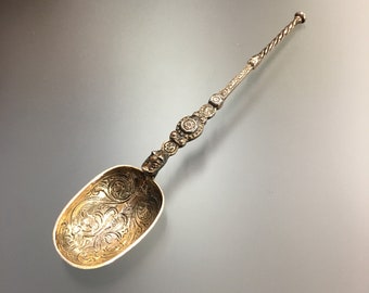 May include: A silver spoon with an ornate handle and a decorative, engraved bowl. The spoon has a long, slender handle with a rounded end.