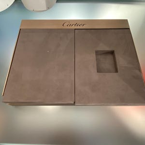 May include: A brown, rectangular, two-piece jewelry box with a gold top. The top of the box has the word 'Cartier' engraved on it. The box is open and the inside is lined with brown velvet. The box has a rectangular cutout in the right side for a ring.