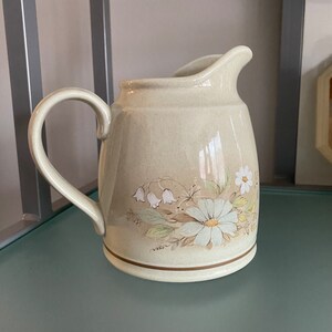 Peut inclure: Pichet en céramique crème avec une anse et un bec courbés. Le pichet est décoré d'un motif floral avec des marguerites blanches et des fleurs en forme de cloche. Une bande marron accentue la base.