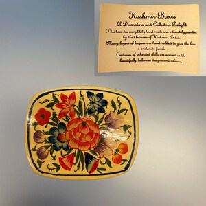 May include: A square, cream-colored box with a hand-painted floral design. The flowers are red, orange, yellow, blue, and purple. The box is decorated with a black outline. "Kashmir Boxes A Decorators and Collectors Delight" is written on a card behind the box.