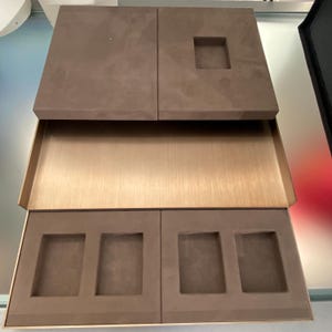May include: A brown suede box with a gold metal tray and four compartments. The box is open and the tray is pulled out, revealing the compartments. The compartments are lined with brown suede and have a rectangular shape.