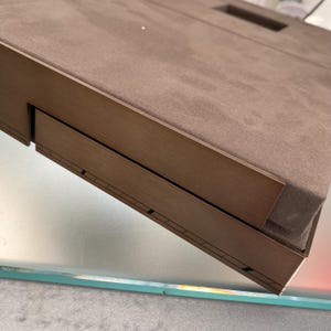 May include: A brown wooden box with a brown suede top and a drawer that is partially open. The box is sitting on a glass surface.