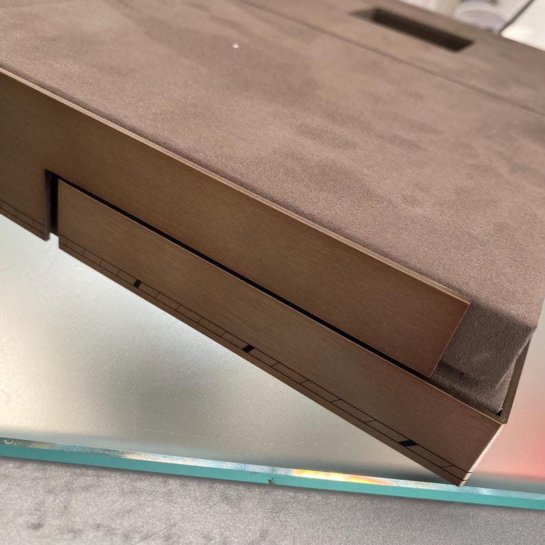 May include: A brown wooden box with a brown suede top and a drawer that is partially open. The box is sitting on a glass surface.