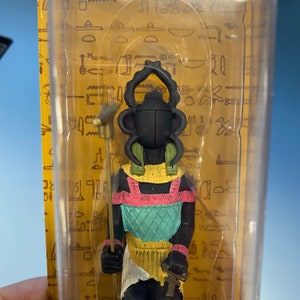 Peut inclure: Une figurine noire et dor&eacute;e d'un dieu &eacute;gyptien avec une t&ecirc;te de scarab&eacute;e, portant une jupe color&eacute;e et tenant un b&acirc;ton. La figurine est pr&eacute;sent&eacute;e dans un bo&icirc;tier en plastique transparent sur fond jaune.