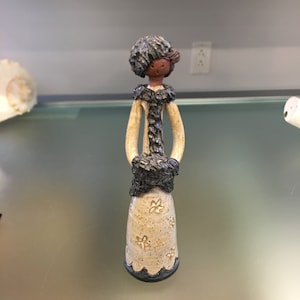 Könnte beinhalten: Eine Keramikfigur einer Frau, die einen braunen Mantel mit schwarzem Pelzkragen und einen schwarzen Hut trägt. Die Figur steht auf einer weißen Oberfläche.