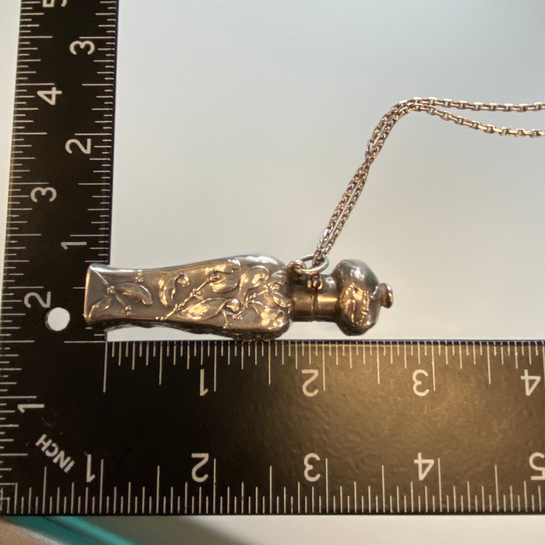 May include: A silver-toned perfume bottle pendant with floral and leaf detailing. The bottle is approximately 2 inches long and hangs from a delicate chain. The bottle has a decorative stopper and is displayed against a light background.