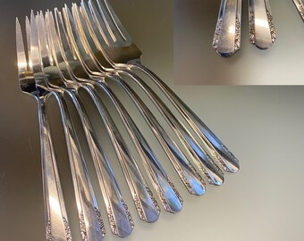 Op de afbeelding: Een set van negen zilveren vorken, in een waaier gerangschikt. Elke vork heeft een decoratief bloemmotief aan de basis van de tanden en langs de steel. De vorken hebben een gepolijste, spiegelachtige afwerking.