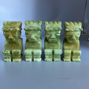 May include: Four green stone sculptures of stylized human figures with square heads and seated poses. The figures have carved facial features and are arranged in a row. The sculptures are a light green color.