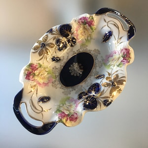 May include: Oval porcelain serving dish with a white base, decorated with pink and yellow floral patterns, gold accents, and navy blue details. The dish has handles and a navy blue oval centre with a gold snowflake design.