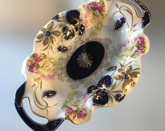 May include: Oval-shaped porcelain serving dish with a white base, featuring pink and yellow floral patterns, gold accents, and navy blue details. The dish has handles and a navy blue oval center with a gold snowflake design.