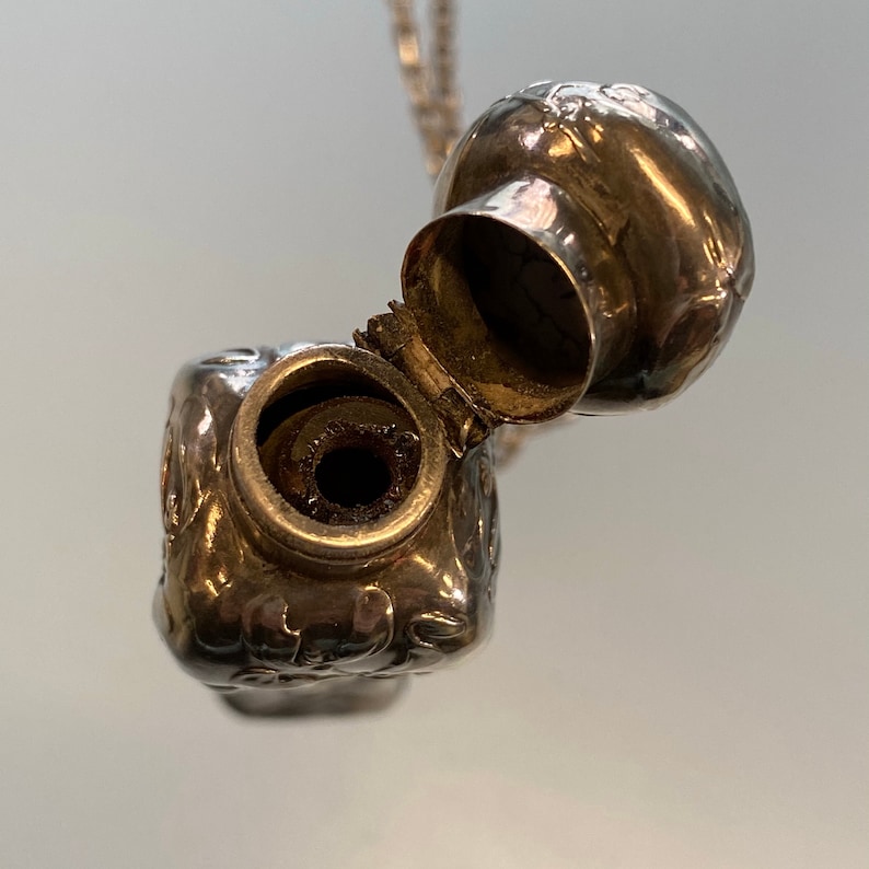 May include: A small, ornate silver pendant with a hinged lid, revealing a dark interior. The pendant is suspended from a delicate silver chain. The surface of the pendant is decorated with embossed floral patterns.