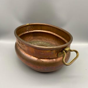 May include: A copper bowl with a brass handle. The bowl has an oval shape and a slightly rounded base. The copper has a reddish-brown color with some areas of darker oxidation. The handle is a light gold color.