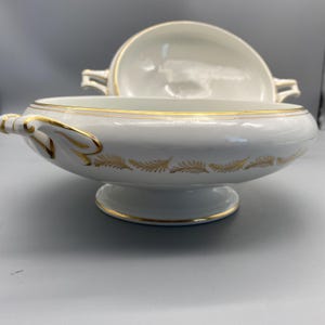 Puede incluir: Una sopera de porcelana blanca con tapa, con ribete dorado y un patrón decorativo de hojas doradas alrededor del cuenco. La sopera tiene asas y se apoya sobre una base de pedestal, mostrando un diseño clásico.