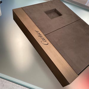 May include: A brown suede jewelry box with a gold trim and the word 'Cartier' engraved on the side.