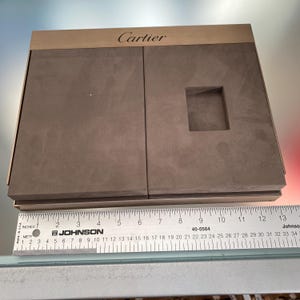 May include: A brown suede Cartier jewelry box with a rectangular cutout for a ring or watch. The box is closed and has a wooden top with the word 'Cartier' printed in gold letters.