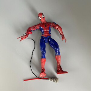 May include: A red and blue action figure of Spider-Man with a web shooter accessory. The figure is posed with one arm outstretched and the other holding the web shooter. The figure is standing on a white surface.