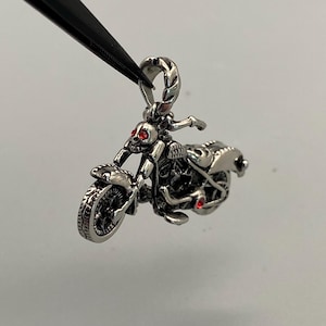 Puede incluir: Colgante de motocicleta plateada con un cráneo como piloto y ojos de gema roja. El colgante está sujeto por una herramienta negra. La motocicleta tiene detalles, incluyendo las ruedas, el asiento y el manillar.