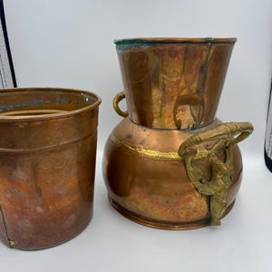 Könnte beinhalten: Zwei Vasen aus Kupfer und Messing mit rundem Korpus und ausgestelltem oberen Teil. Die größere Vase hat einen dekorativen Griff mit einem Vogelmotiv.