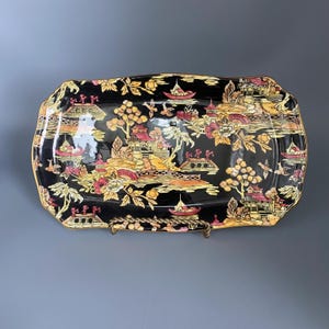 Peut inclure: Plateau de service rectangulaire en céramique noire avec un bord doré. Le plateau présente un motif de paysage d'inspiration asiatique détaillé dans des tons de rouge, jaune et or. Le motif comprend des bateaux, des ponts et des bâtiments.