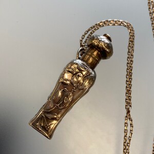 May include: A silver-toned perfume bottle pendant with a decorative floral and face design. The bottle is suspended from a delicate chain. The bottle's design features a bulbous top and a slender, elongated body.