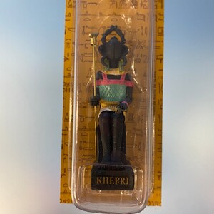 Peut inclure: Une petite figurine noire du dieu &eacute;gyptien Kh&eacute;pri, repr&eacute;sent&eacute; comme un scarab&eacute;e avec une t&ecirc;te humaine, portant une coiffe color&eacute;e et tenant un b&acirc;ton. La figurine est assise sur un tr&ocirc;ne et est expos&eacute;e dans un bo&icirc;tier en plastique transparent sur fond jaune. Le bo&icirc;tier porte l'inscription "KHEPRI" en bas.