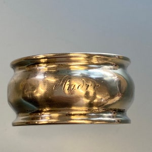 May include: Silver napkin ring with a rounded shape and a raised edge. The ring is engraved with the word "Andre".