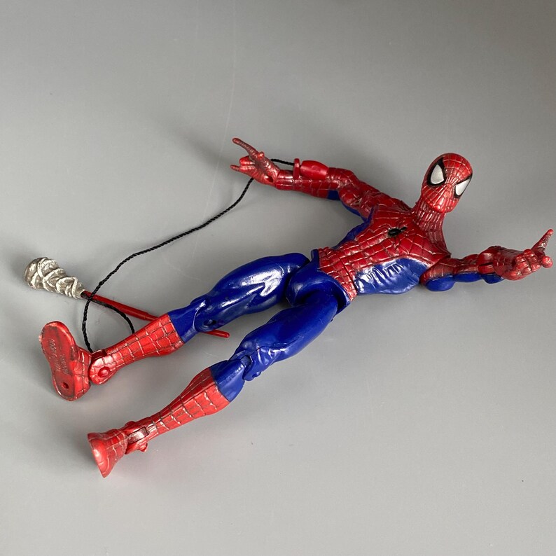 May include: A red and blue action figure of Spider-Man with a web shooter accessory. The figure is lying on its back with its arms outstretched.