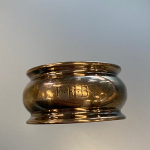 May include: A silver napkin ring with a rounded shape and a raised rim. The ring is engraved with the initials "F.H.B."