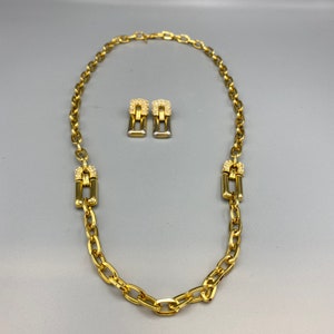 Può includere: Una collana a catena d'oro con un designo a maglie e due orecchini d'oro abbinati con pietre trasparenti. Gli orecchini hanno la forma di un rettangolo con un piccolo quadrato in cima.