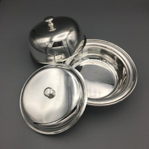 Puede incluir: Tres brillantes fuentes de plata con tapas. Una tapa está abierta, revelando el interior del plato. La otra tapa está parcialmente abierta y la tercera está cerrada. Los platos están sobre una superficie gris.