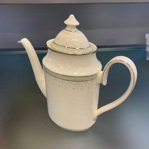 Puede incluir: Una cafetera de cerámica blanca con tapa y ribete verde y dorado. La cafetera tiene un delicado patrón floral.